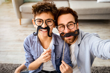 Funny selfie. Happy father and son taking selfie, holding fake moustache on sticks, wearing glasses...