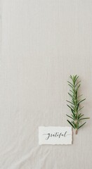A clean minimalist top-down composition displays a small white card with the word "grateful" next to a fresh rosemary sprig on a light textured fabric.