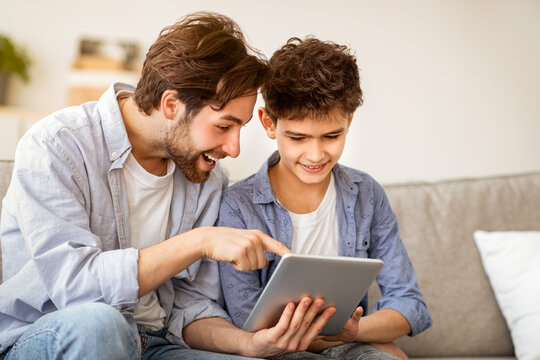 Young father teaching his son to use new application on digital tablet, surfing internet while spending time together at home, copy space. Leisure activities, modern technologies concept