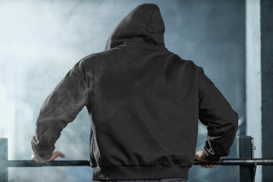 In a gym environment a man wearing a black hooded sweatshirt positions himself by a bench press barbell ready to engage in a strength training session focused on muscle building and fitness goals.