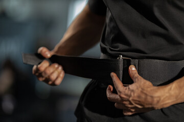 man in a black shirt adjusts his squat belt while getting ready to lift a barbell in a gym. He appears focused and ready for an intense workout in the fitness center.