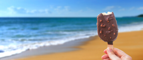 Chocolate covered ice cream bar in the summer at the beach, during the holidays. Hand holds delicious chocolate and nuts almond ice cream bar on a stick.