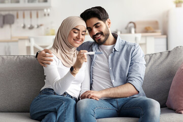 Obraz premium Positive emotion, joyful couple, become parents, happy news, expect for baby. Smiling arab millennial husband and wife hugs rejoice to pregnancy test result, sits on sofa in home interior, free space