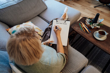 Middle aged woman writing New year's goals on note paper. New year resolutions list, cup of tea on...