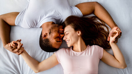 Top view of young multiracial couple lying on bed, cuddling, expressing affection to each other....