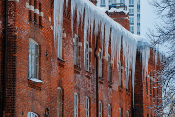 Long, sharp icicles are hanging from the snow-covered roof of an old brick building, creating a...