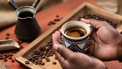 A pair of hands holds a traditional small ceramic cup filled with a dark, hot beverage, likely coffee. An old metal coffee pot and scattered coffee beans on a wooden tray.