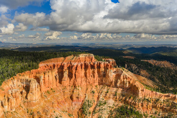 Fototapeta premium Dramatic red rock cliffs and hoodoos at Cedar Breaks National Monument in Utah, with forested mountains and clouds under a bright blue sky.