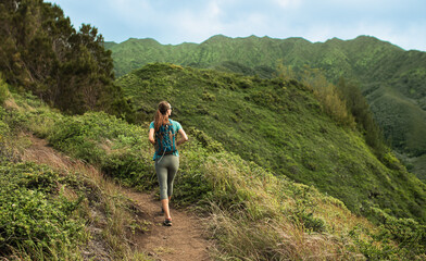Fototapeta premium A woman with a backpack hiking along a picturesque green Hawaii hills trail under blue skies surrounded by lush nature. 
