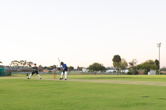 Male teenage wicketkeeper crouching behind stumps and batsman in helmet on pitch holding bat
