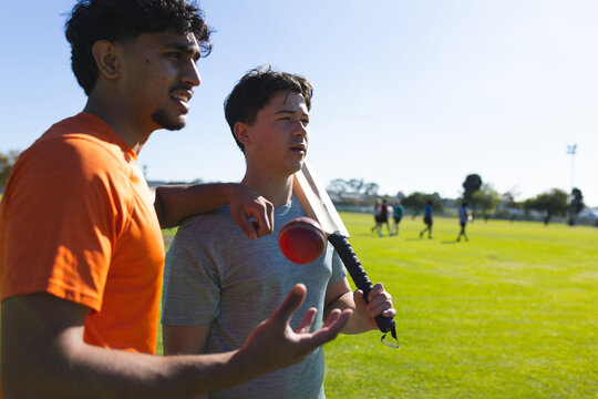 Diverse male teammates discussing batting technique on grass field with cricket bat and ball