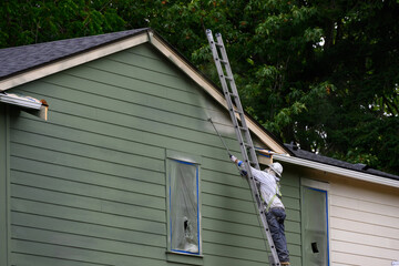 Professional painter wearing a hard hat and respirator on a ladder using a sprayer to paint the exterior of a new build house green, new housing development under construction  © knelson20