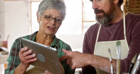 Obraz premium Pointing at screen coworkers holding tablet at wooden table in art studio with paintbrushes, aprons