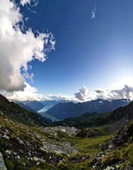 Fototapeta premium A vast mountain vista reveals a valley nestled amongst alpine peaks, with scattered clouds painting the vibrant blue sky.