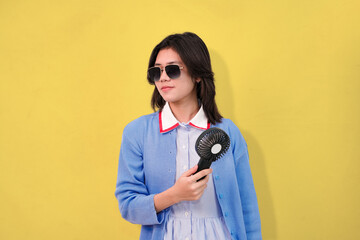 A confident young woman cooling herself with a hand-held fan while wearing sunglasses