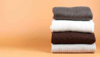 A stack of folded sweaters in neutral tones, including gray, white, and brown, displayed against a soft peach background