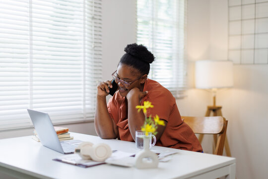 Happy positive woman entrepreneur sitting in home office, working on laptop computer, talking on cell phone.
