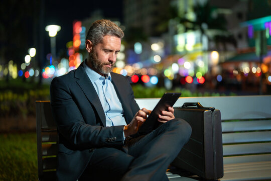 Businessman working on tablet outdoors. Businessman with tablet outdoor. Businessman with a tablet on night street. Businessman using tablet, sit on bench. Night business.
