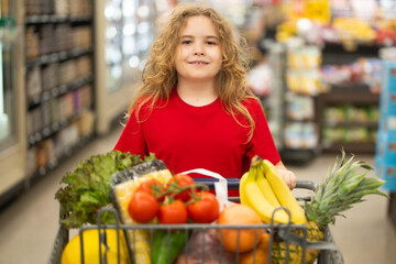 Child shopping with cart in supermarket. Kid choosing fresh vegetables in grocery store. Child with food in supermarket. Kid selecting produce at supermarket. Child with fresh food in supermarket.
