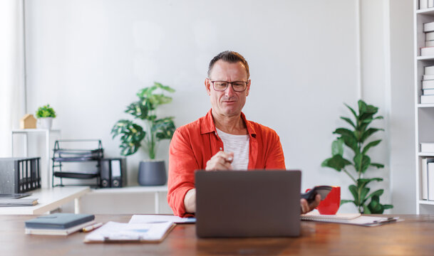 Serious and focused financial accounting on paper work at office, mature man using calculator and laptop for calculating reports and summarizing accounts, businessman at work in casual clothes.