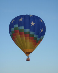 Hot air balloon floating against a clear blue sky. Vibrant blue envelope with stars and rainbow bands, clean negative space for copy, and uplifting travel mood.
