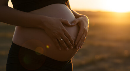 A pregnant woman's belly, adorned with a heart shape formed by her hands, bathed in the warm glow of a sunset outdoors.