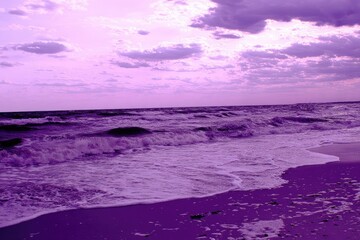 Obraz premium Purple-hued ocean waves lapping a sandy shore under a cloudy sky