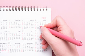 Calendar and female hand with pen on pink background close-up top view.