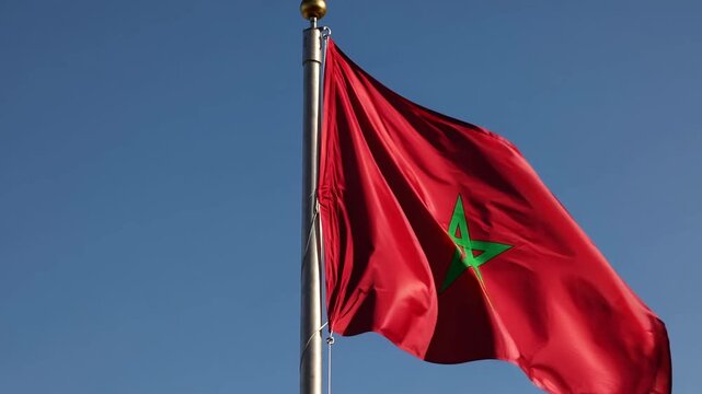 Low-angle video shot of a Moroccan flag waving against a clear blue sky, capturing the vibrant red and green colors in a dynamic, patriotic style.