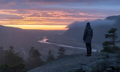 Person views a river valley at sunrise from a rocky outcrop, mist in distance