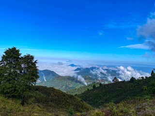Stunning mountain landscape with green hills, trees, and clouds covering the valley under a clear blue sky.