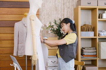 Young asian woman using both hands to press fabric on mannequin during garment fitting inside home fashion studio with shelves full of folded fabric, boxes and handmade sewing tools for design work
