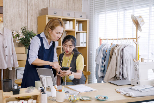 Young asian fashion designer and senior female partner reviewing smartphone content during business planning session in home studio workspace surrounded by sewing equipment and clothing samples