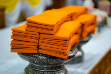 Close-up of a stack of orange monk's robes, neatly folded on an ornate silver pedestal, with other elements in the background.