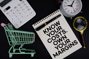 Know your costs. Own your margins text written on notepad with alarm clock and trolley shopping...