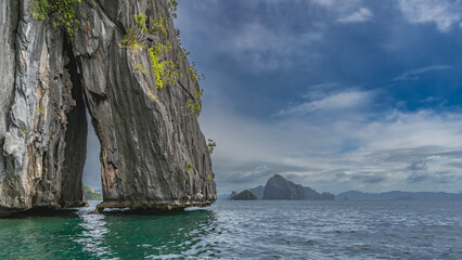 A picturesque cliff above the ocean. The gap between the steep slopes. Green vegetation on the rock. Mountains on the horizon. Blue sky, clouds. Philippines. Palawan. El Nido.