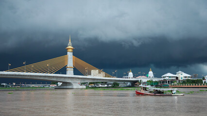 Cable-stayed bridge with Thai-style golden spire over river under dramatic stormy sky, with...
