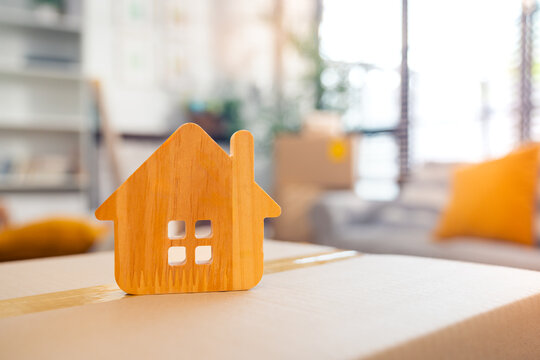 Wooden house icon on cardboard box in modern home interior, symbolizing moving house, new home ownership, real estate purchase, relocation, or first-time buyer lifestyle concept.