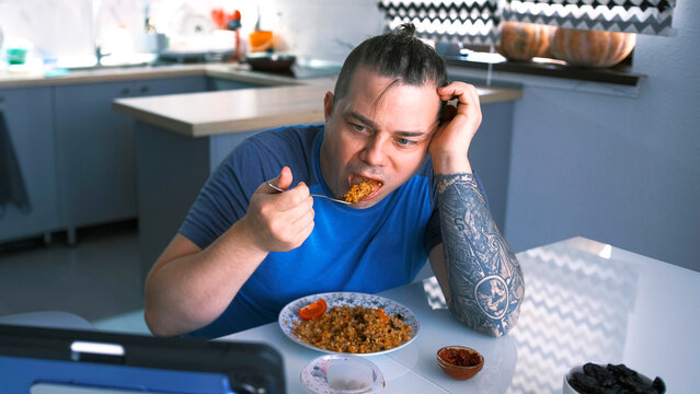 A fat man is eating in the kitchen. A funny guy eats fried bulgur with vegetables while watching a movie on his tablet