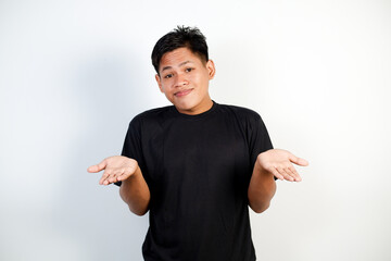 Portrait of a young Asian man in black shirt shrugging with a confused face against a white...