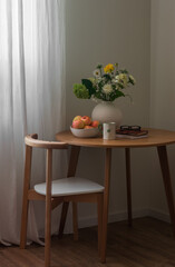 The cozy interior of the living room - a bouquet of flowers in a ceramic vase, a bowl of apples, books, and a glass of tea on a round wooden table