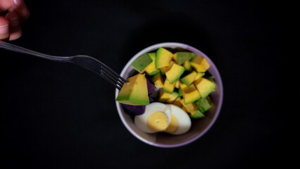Fork piercing healthy avocado and boiled egg salad in a bowl, top view, dark background