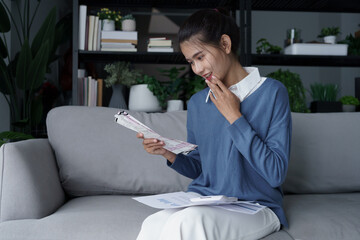 Asian woman sits on sofa using calculator calculating monthly expenses in living room