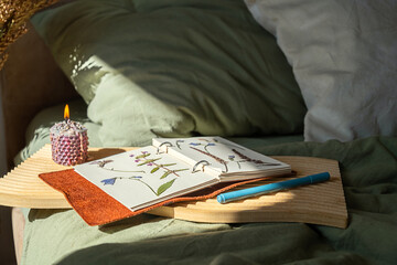 Dried flowers pressed in notebook with burning candle on wooden tray on the bed.
