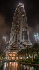A modern architectural , a tall, stylized building stands illuminated at night, reflecting in the tranquil water of a surrounding pool.