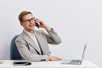 Professional smiling man in formal attire talking on mobile phone while seated at desk with laptop isolated on plain white background studio portrait. Business workplace concept