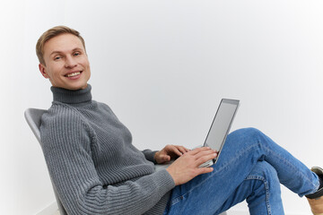 smiling young man working on laptop casual gray sweater blue jeans sitting relaxed white background...