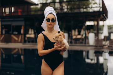 Young woman wearing towel turban and sunglasses holding puppy by poolside