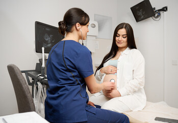 Female doctor listening to pregnant woman's belly with stethoscope during prenatal checkup. Medical...