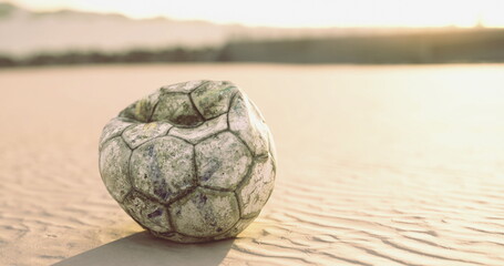 Fototapeta premium A weathered soccer ball lies abandoned on the sandy beach, illuminated by the warm colors of sunset. The sun casts a soft glow, creating a serene atmosphere around the forgotten toy.